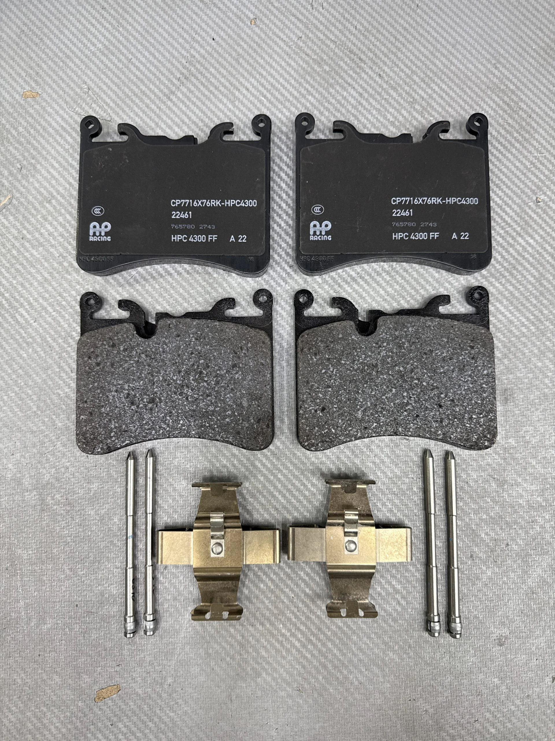 Set of four motorcycle/car brake pads with metal clips and pins laid out on a gray textured surface; two large black pads labeled HPC 4300 FF, and two worn, roughened pads beneath them.