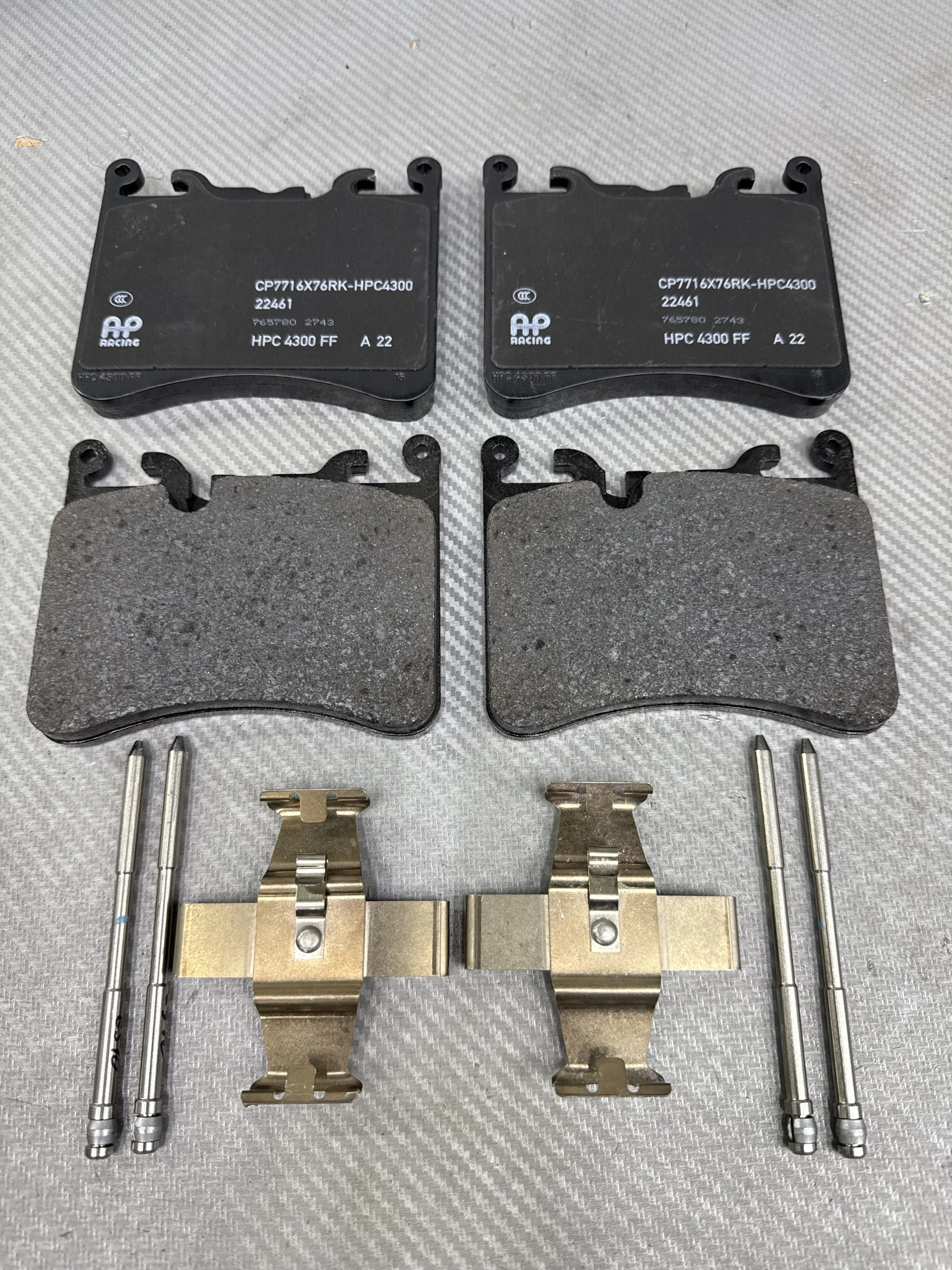Set of four automotive brake pads with accompanying metal hardware laid on a gray textured surface (shows two larger rear pads and two smaller paired pads).