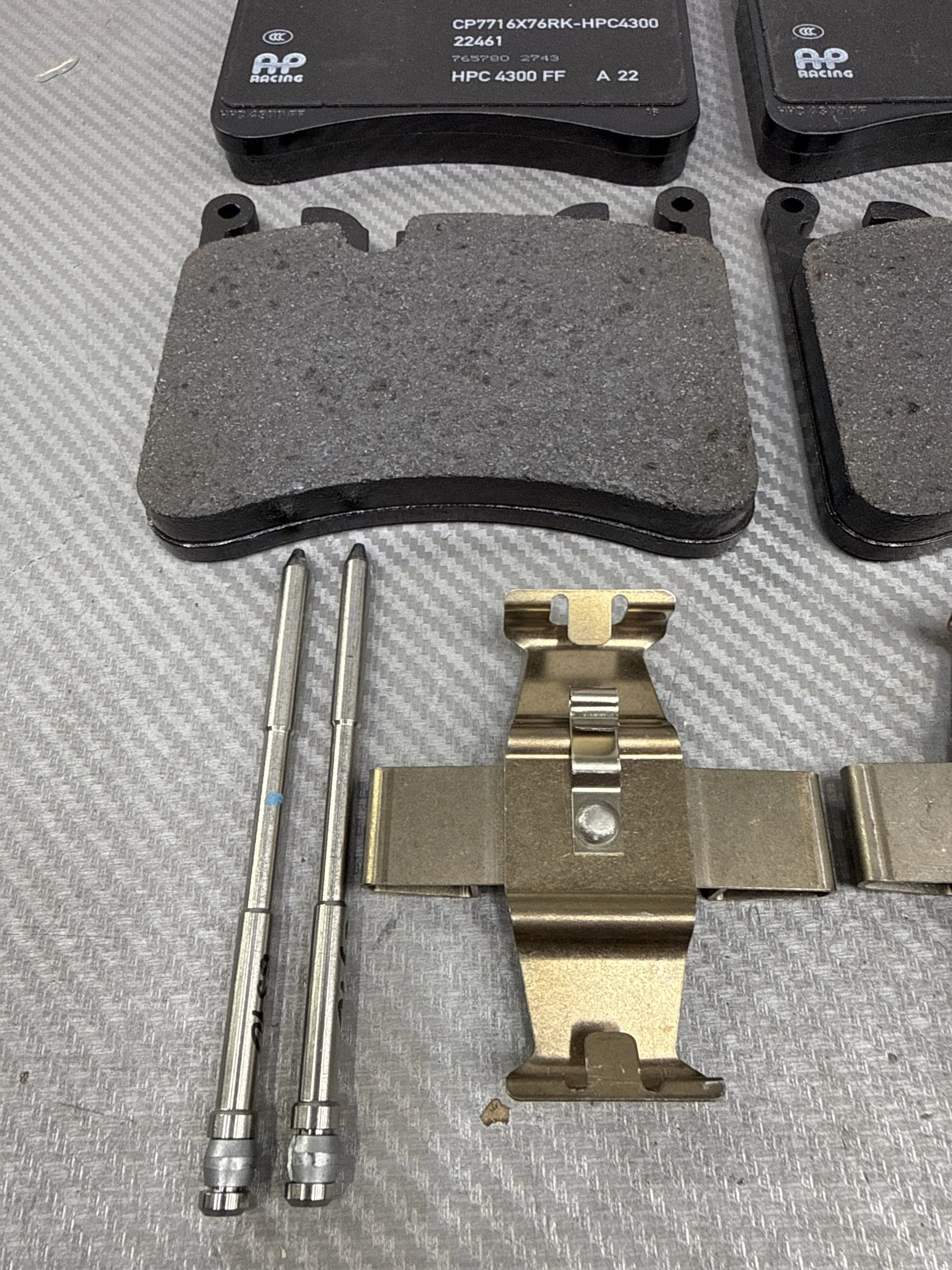 Two black brake pads with gray friction material, alongside metal pins and a brake caliper bracket on a textured work surface.