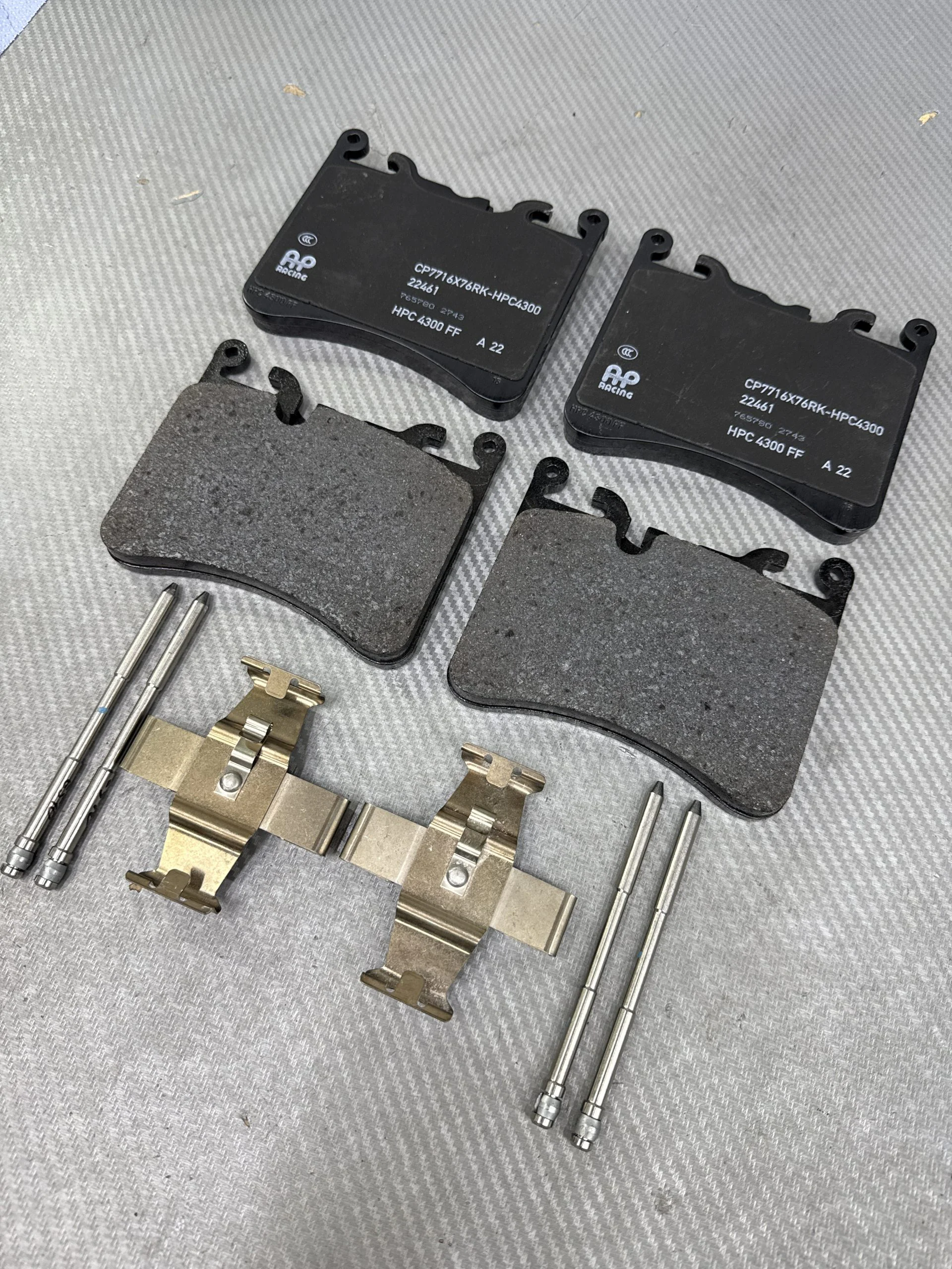 Pair of black brake pads with RP Racing branding, along with metal pins, clips, and shims laid out on a gray textured surface.