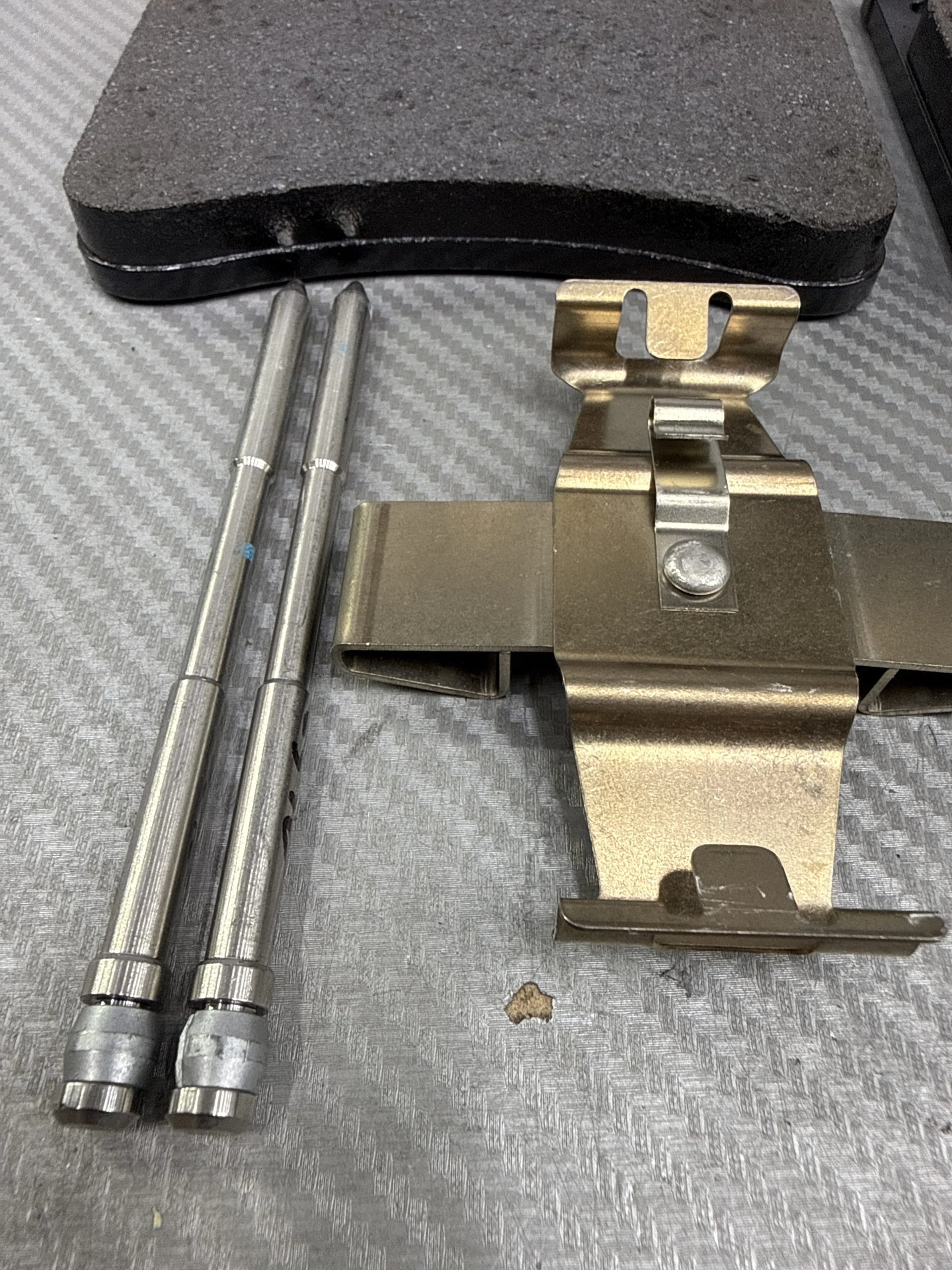 Close-up of a metal mounting bracket with a hinge, two silver pins, and a black foam pad on a textured floor surface.