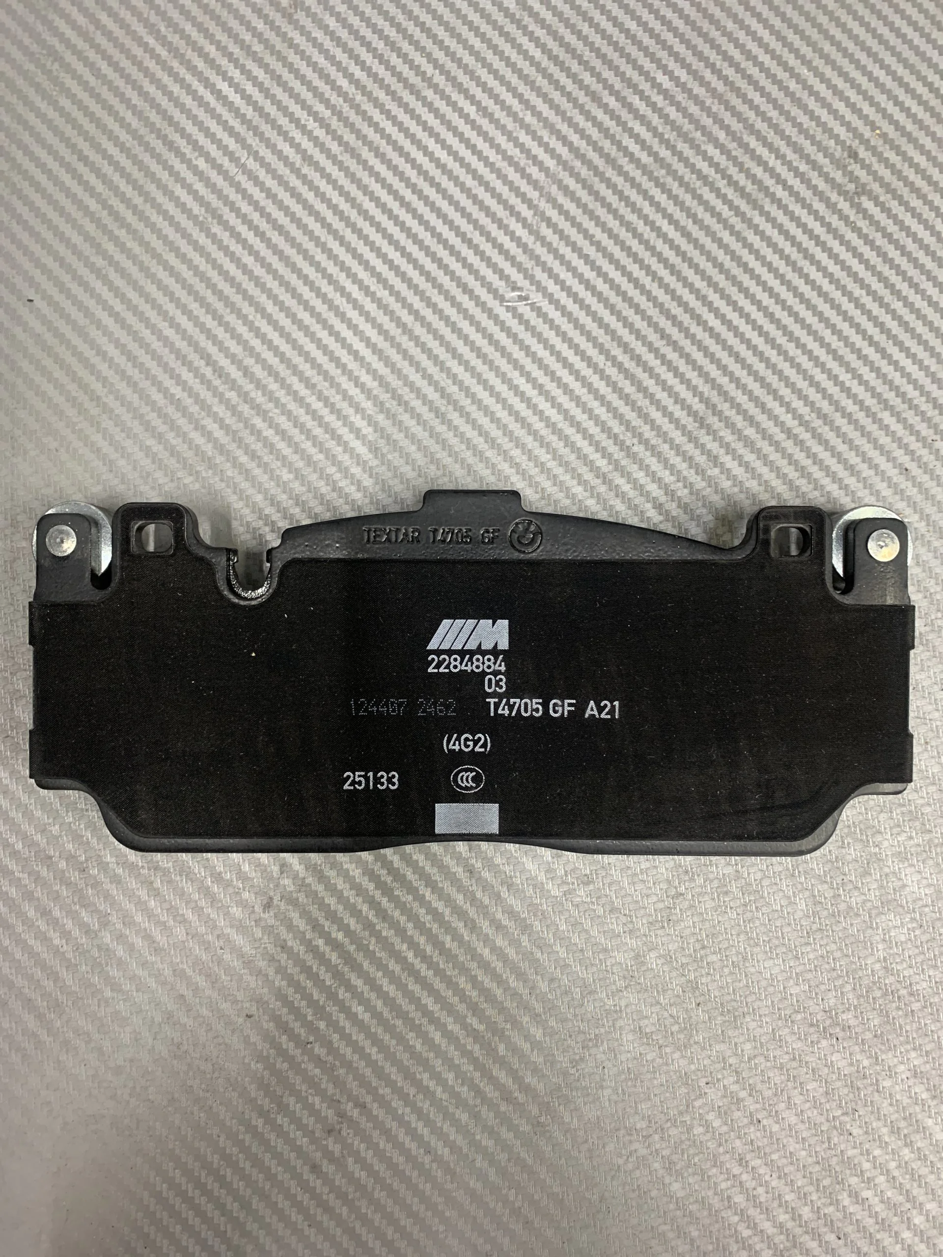 Black automotive control module with the BMW M logo and serial numbers resting on a light gray, textured surface with metal mounting lugs at each end.