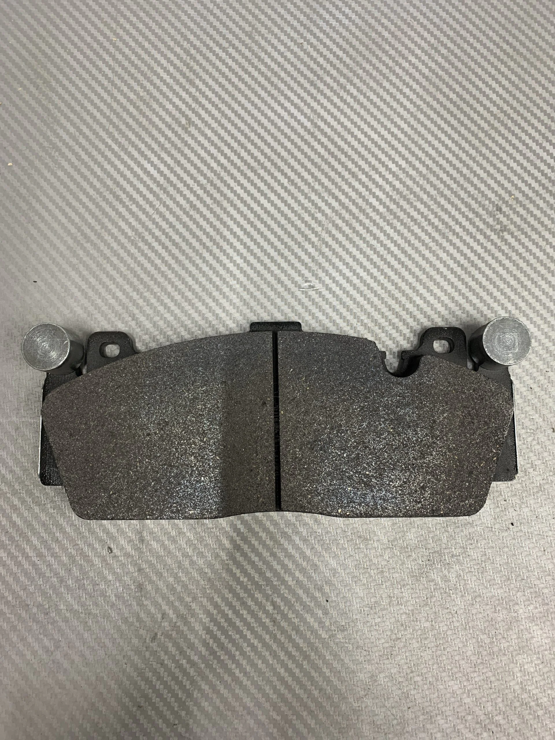 Two dark gray brake pads placed side by side on a textured gray surface, with small metal pins visible at their outer ends.