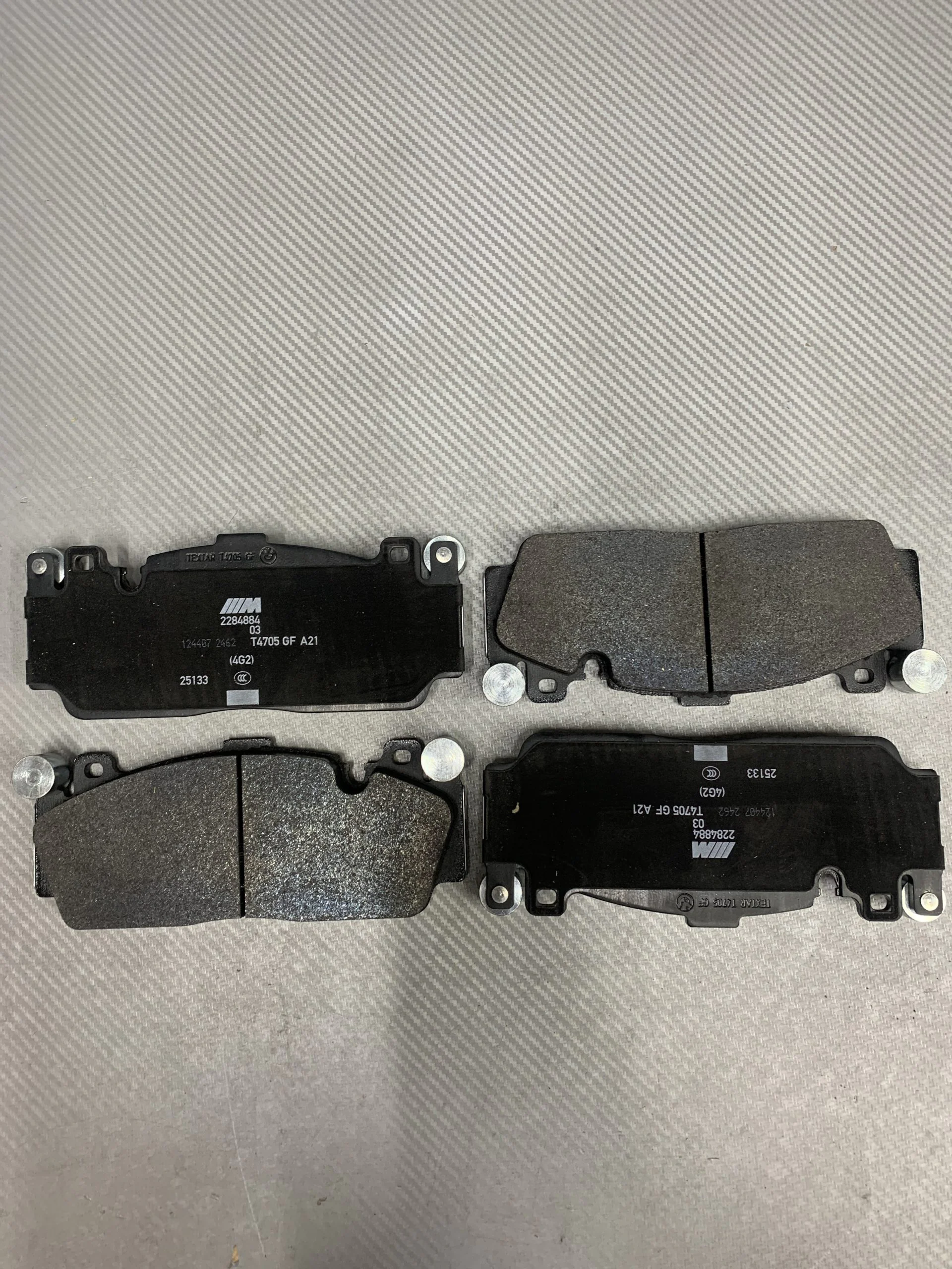 Row of black automotive brake pads laid out on a light gray textured surface, showing friction material and metal mounting points