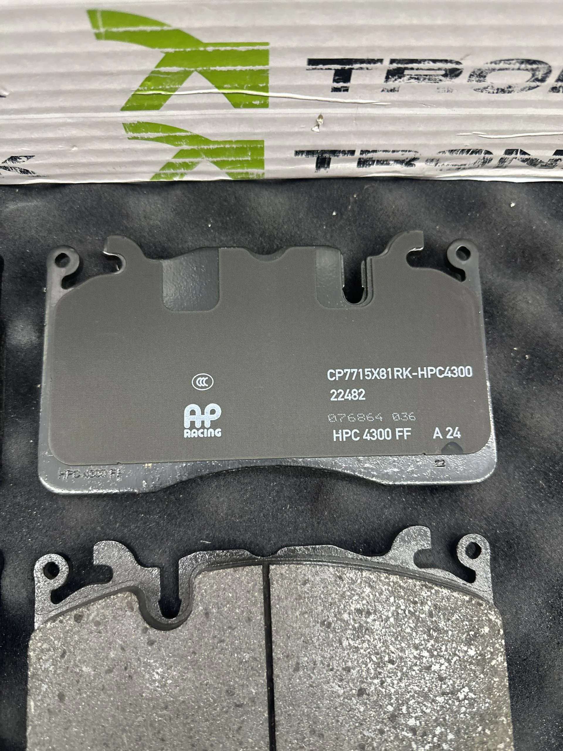 Close-up of a grey AP Racing brake pad with cutouts, resting on a dark surface with packaging above showing the logo.