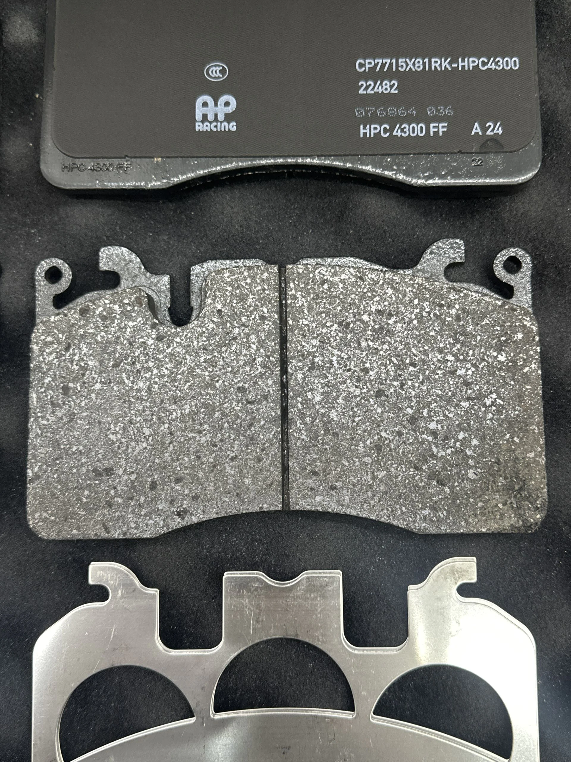 Close-up of a gray brake pad with a worn metal backing plate, showing AP Racing branding and part numbers HPC 4300 FF on a dark surface.