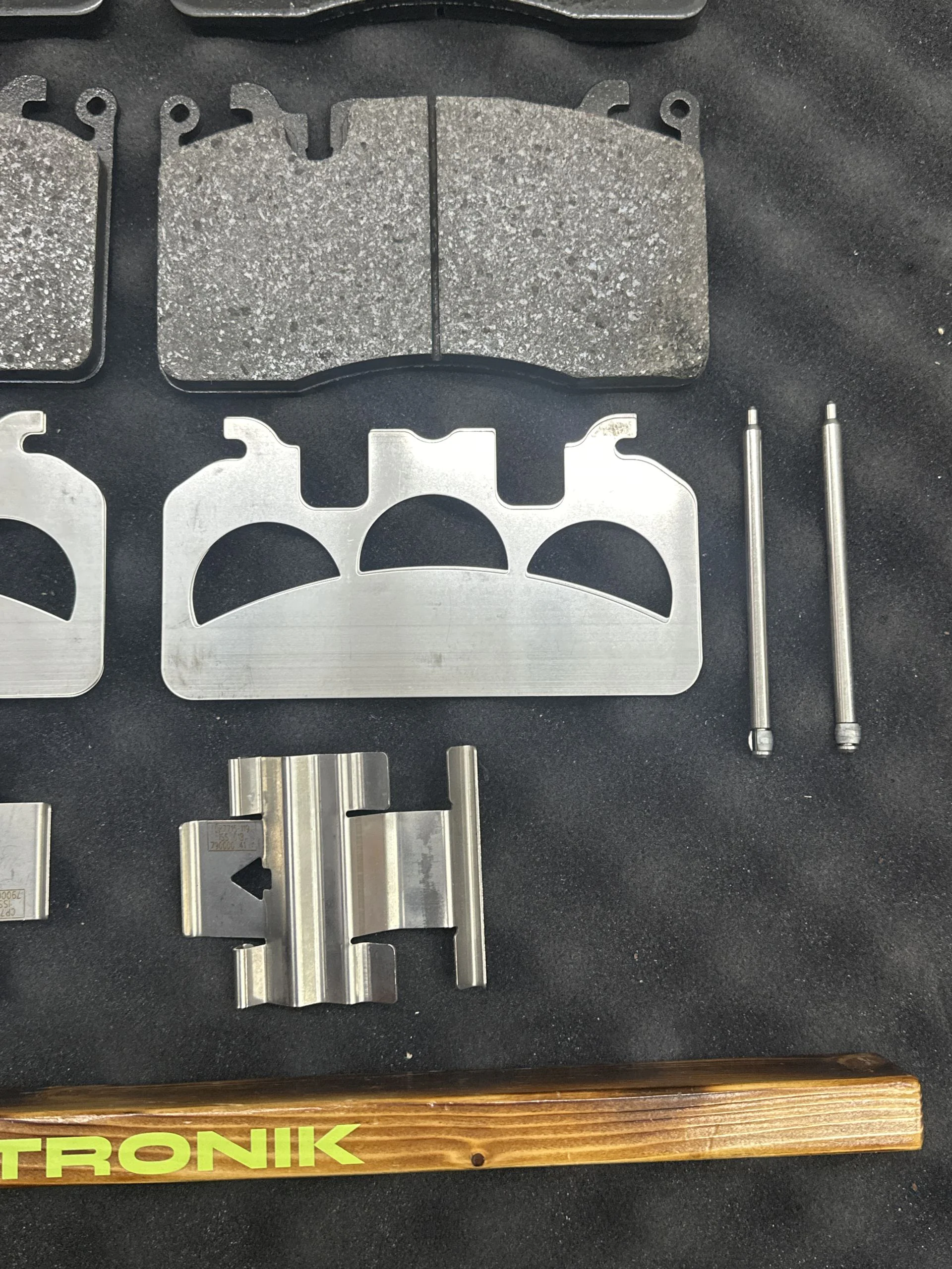 Set of automotive brake pads and metal shims laid out on a dark background, with a wooden ruler showing green text at the bottom for scale