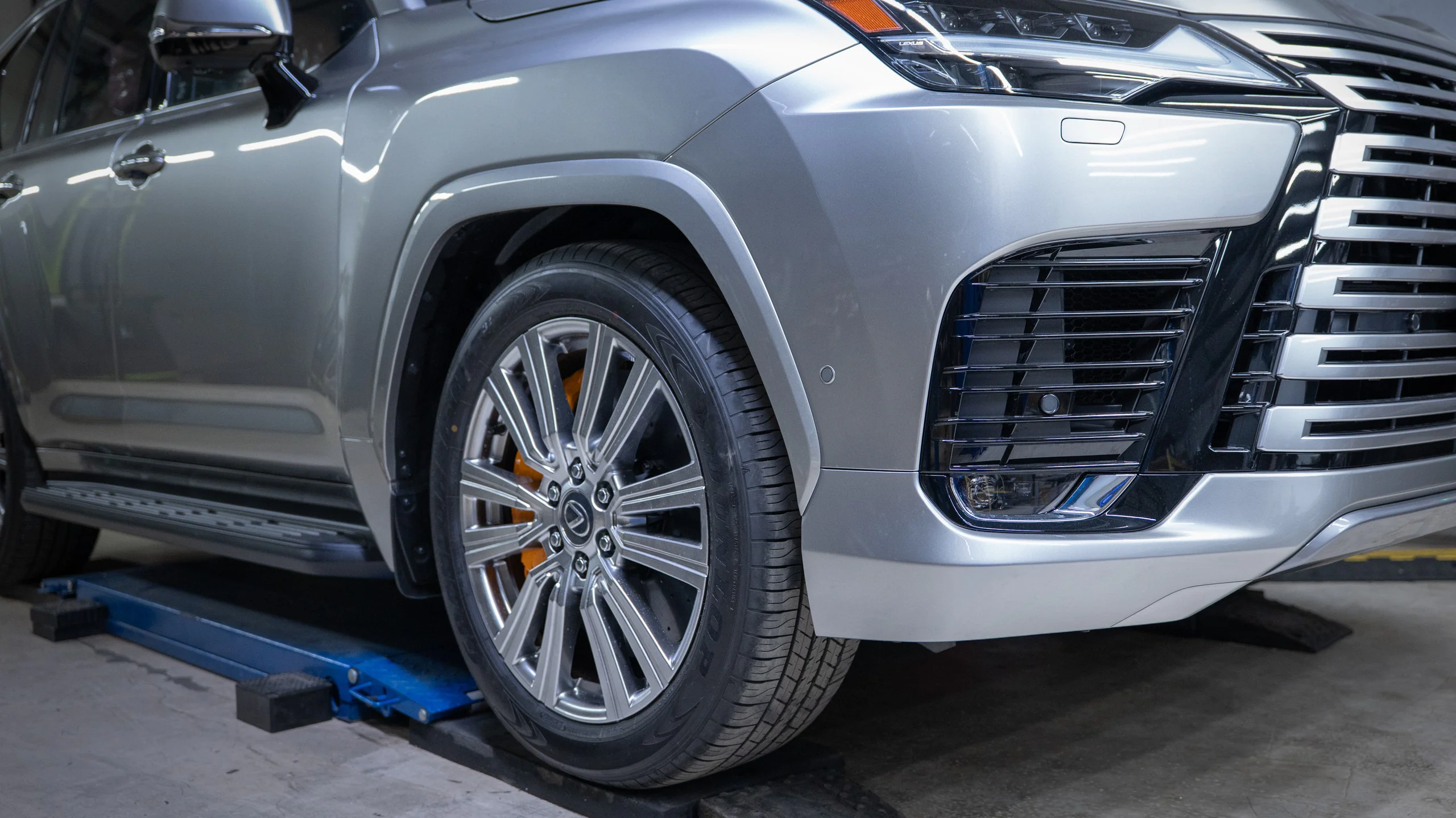 Close-up of a silver SUV on a car lift, showing the front grille, headlight area, and alloy wheel with orange brake calipers in a workshop.